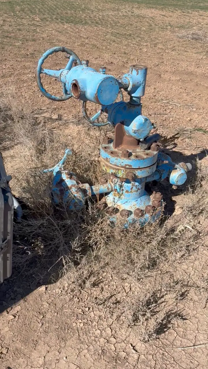 A blue-painted orphaned wellhead with its valve wheel, standing in dry, cracked farm ground