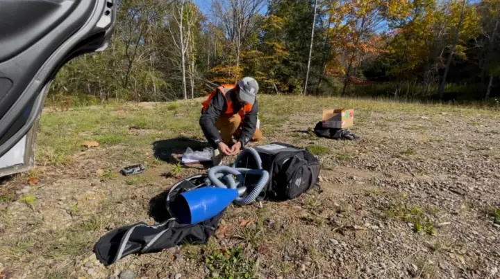 A surveyor in a high-visibility vest kneeling on a gravel clearing to unpack a Hi-Flow sampler, its hoses and blue capture hood on the ground