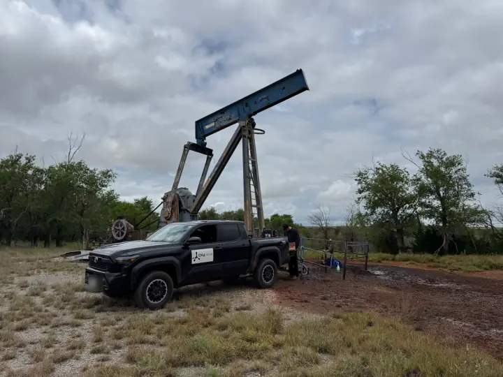 A CH4mber pickup parked beside an old pumpjack on a fenced pad, trees behind under a cloudy sky