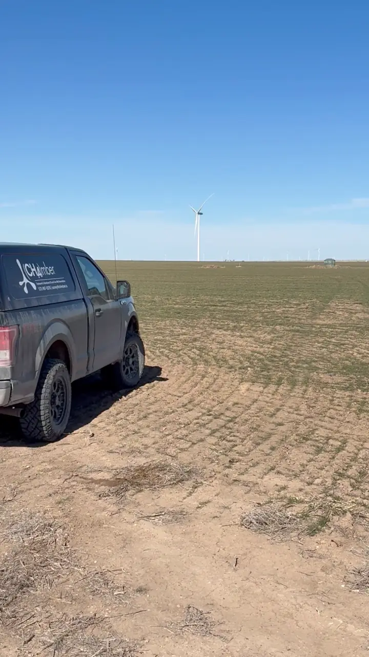 A CH4mber pickup parked at the edge of a bare, worked field running to wind turbines on the horizon