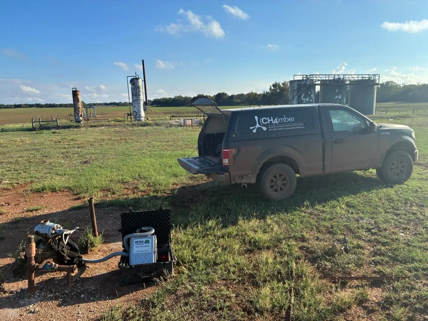A Hi-Flow sampler in its open case beside a rusted wellhead, with a survey truck parked in the pasture behind it