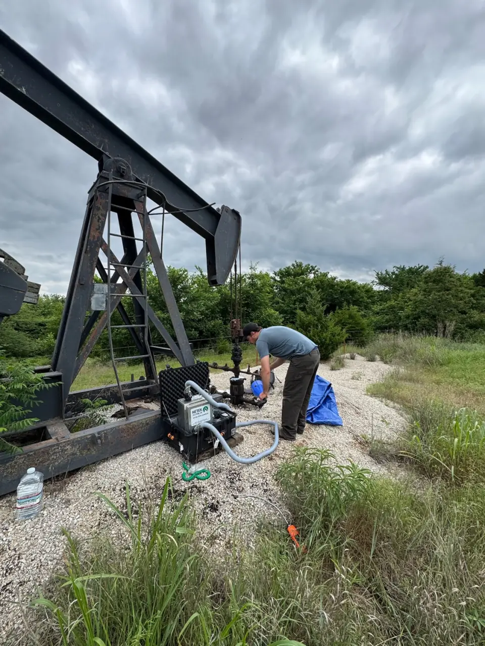 A surveyor bent over a Hi-Flow sampler in an open case on a gravel pad beneath an old pumpjack, hoses running to the wellhead
