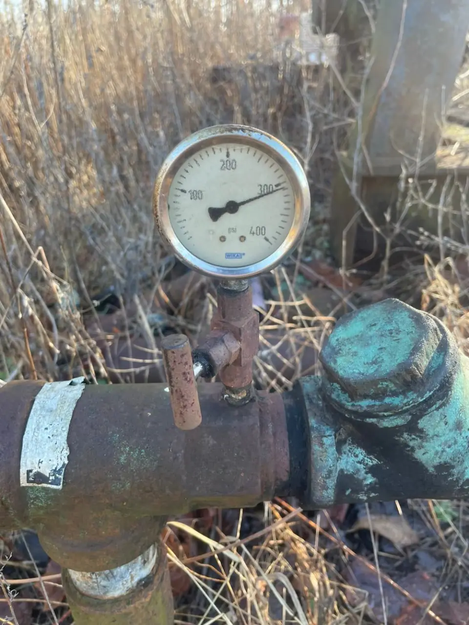 A pressure gauge on a rusted, paint-flaked orphaned wellhead, standing in dry grass