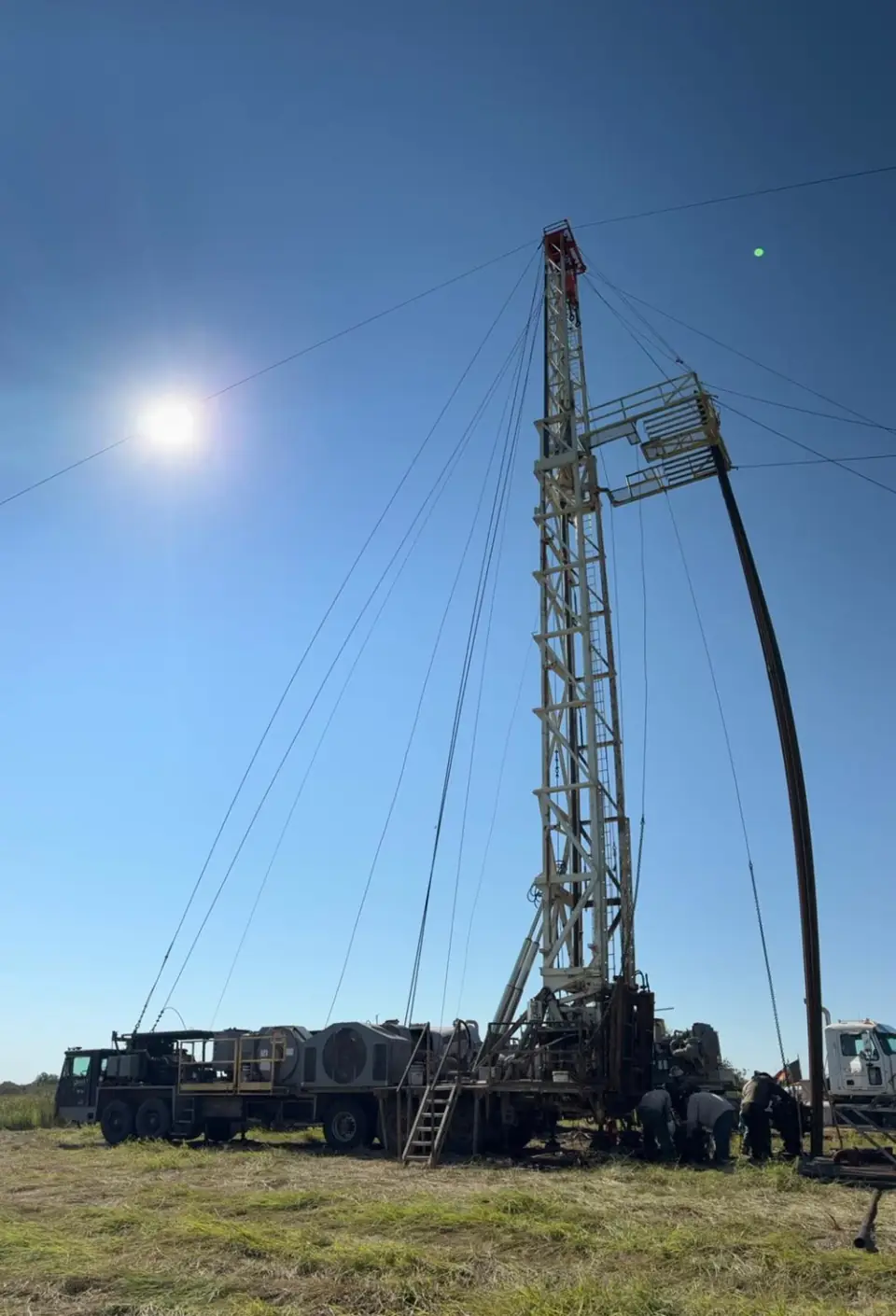 A plugging rig's mast raised over an orphaned well in a grass pasture under a low sun, with the crew working at its base