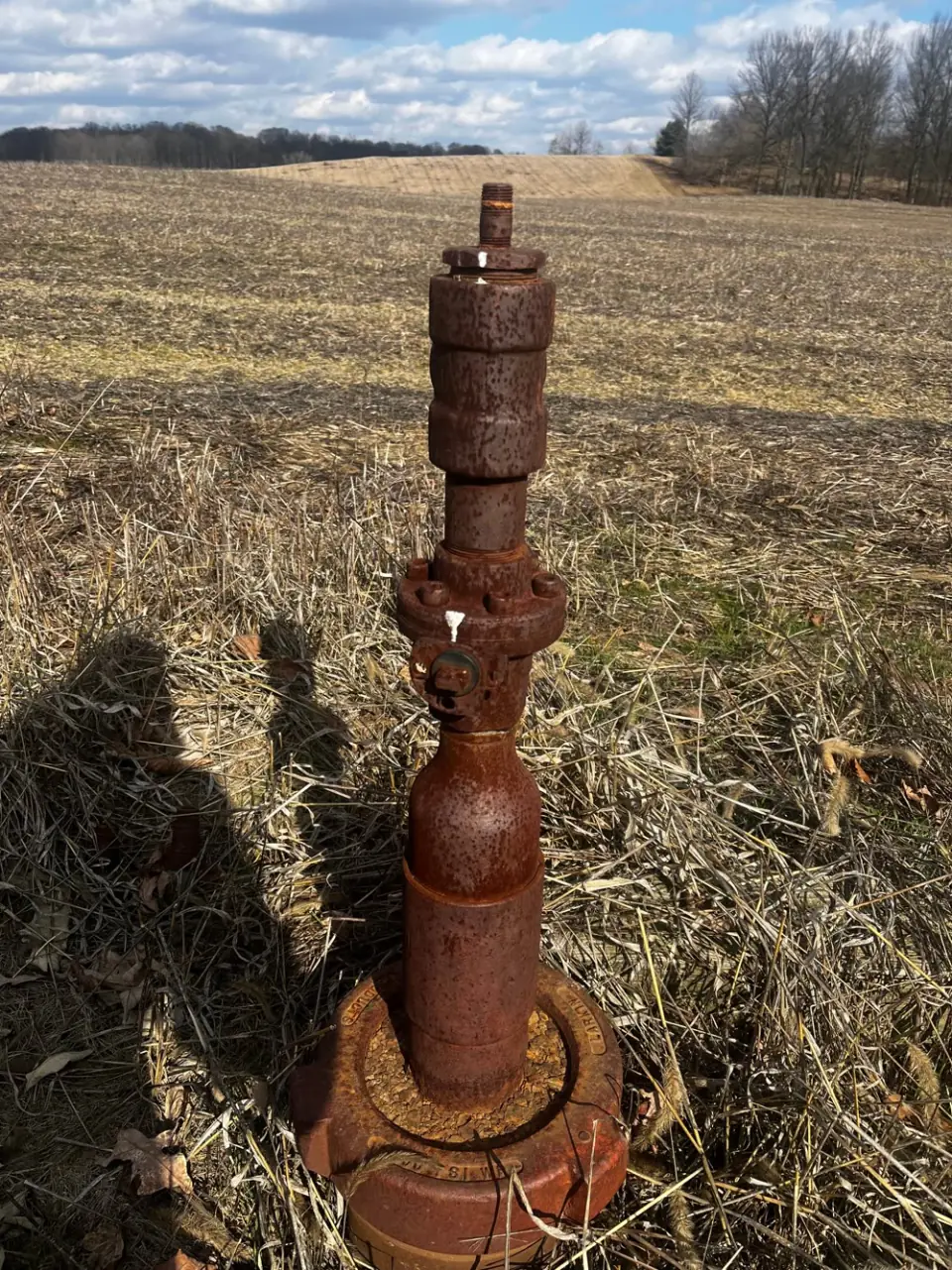 A rusted orphaned wellhead standing in a harvested field, with the shadows of two people beside it